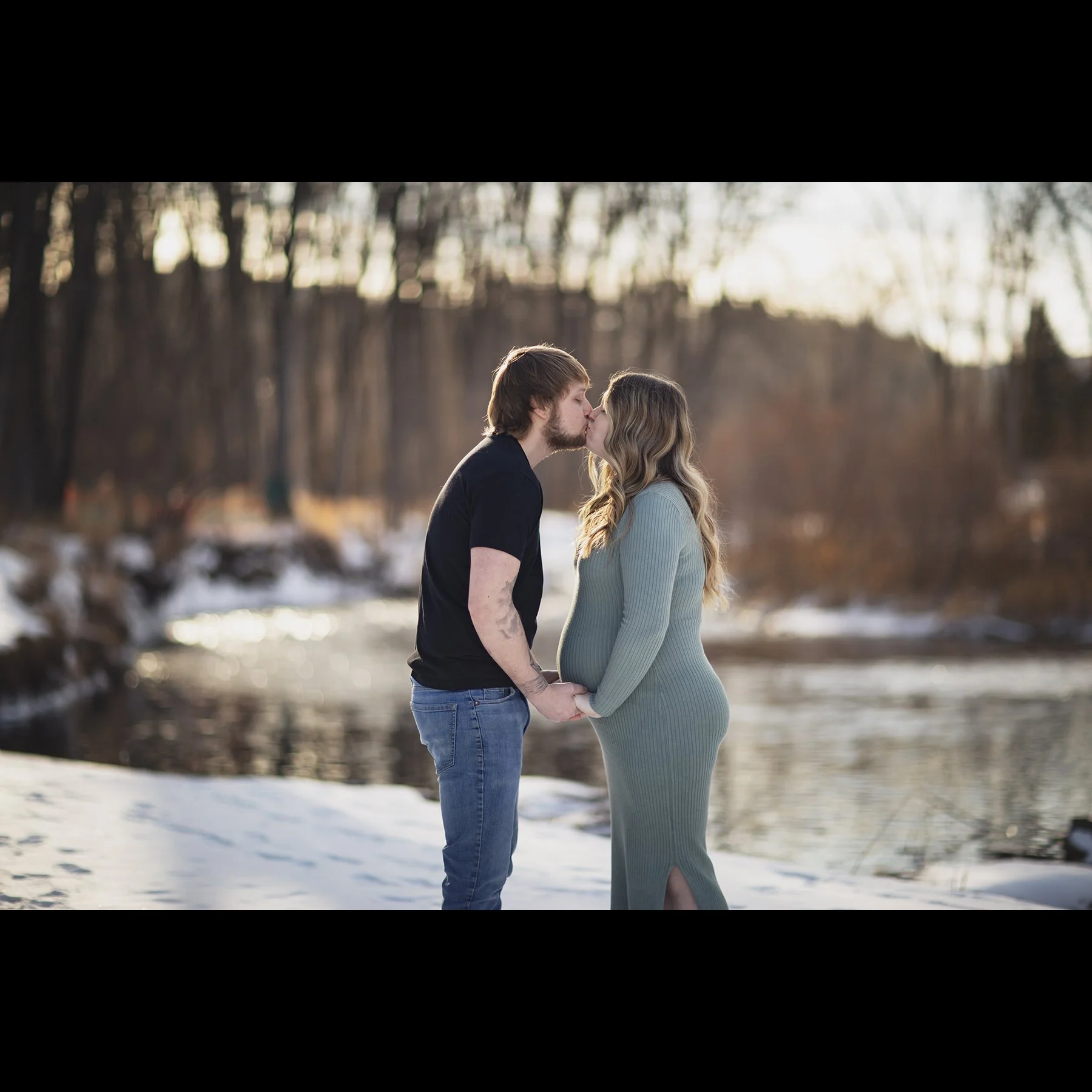 A couple Kissing by a winter river, holding hands, with snow on the ground and leafless trees in the background.