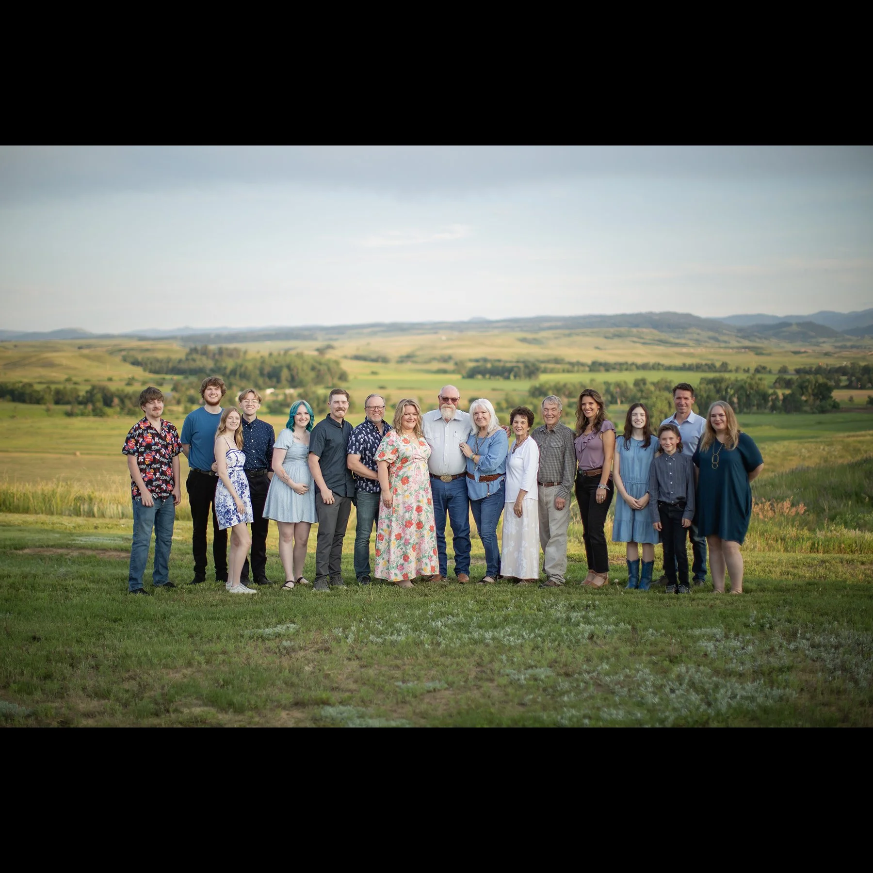 Family group photo outdoors on a grassy field with rolling hills in the background, featuring adults and children smiling and standing together.