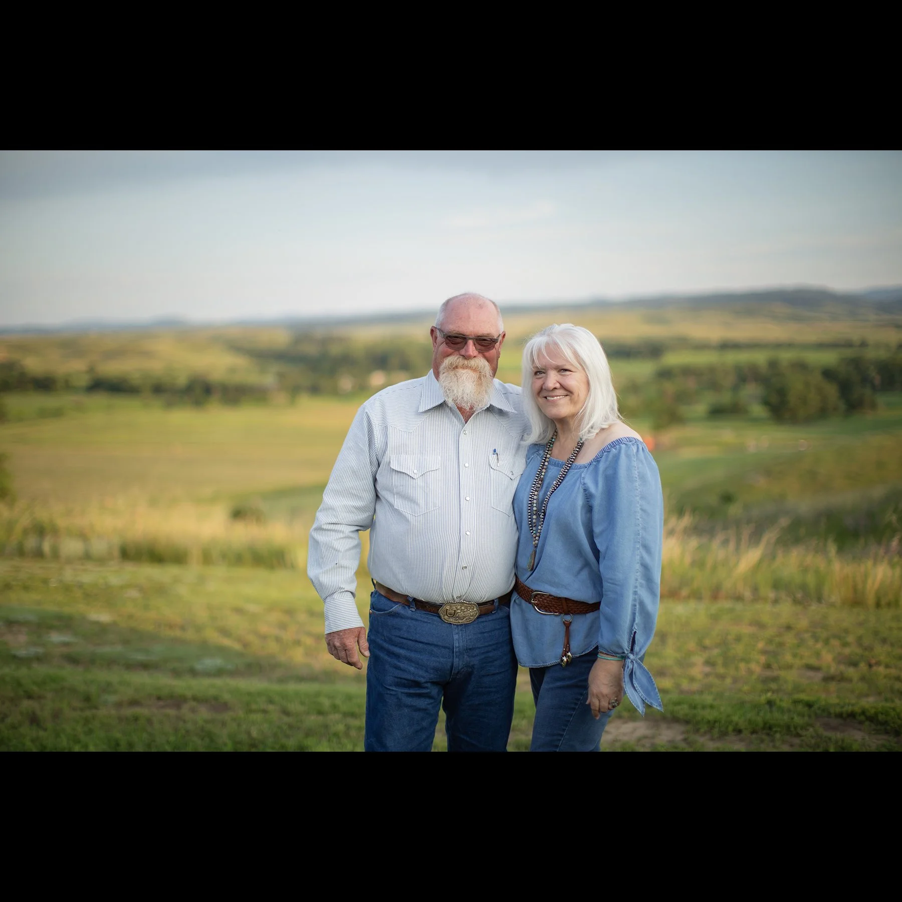 An elderly couple standing outdoors in a scenic field with rolling hills in the background, smiling at the camera.