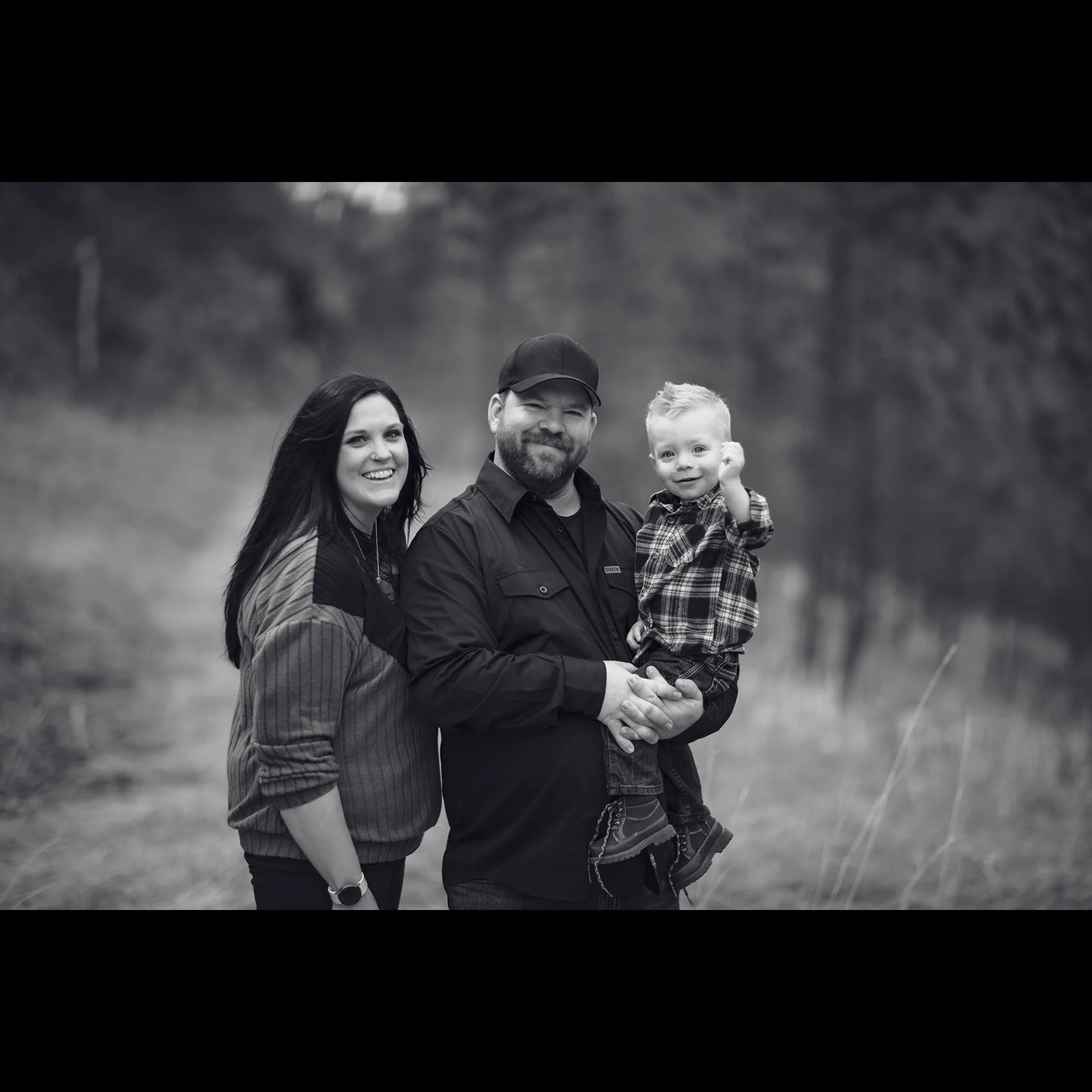 Black and white photo of a smiling family outdoors, with a woman, a man holding a young boy, standing in a grassy area with blurred trees in the background.