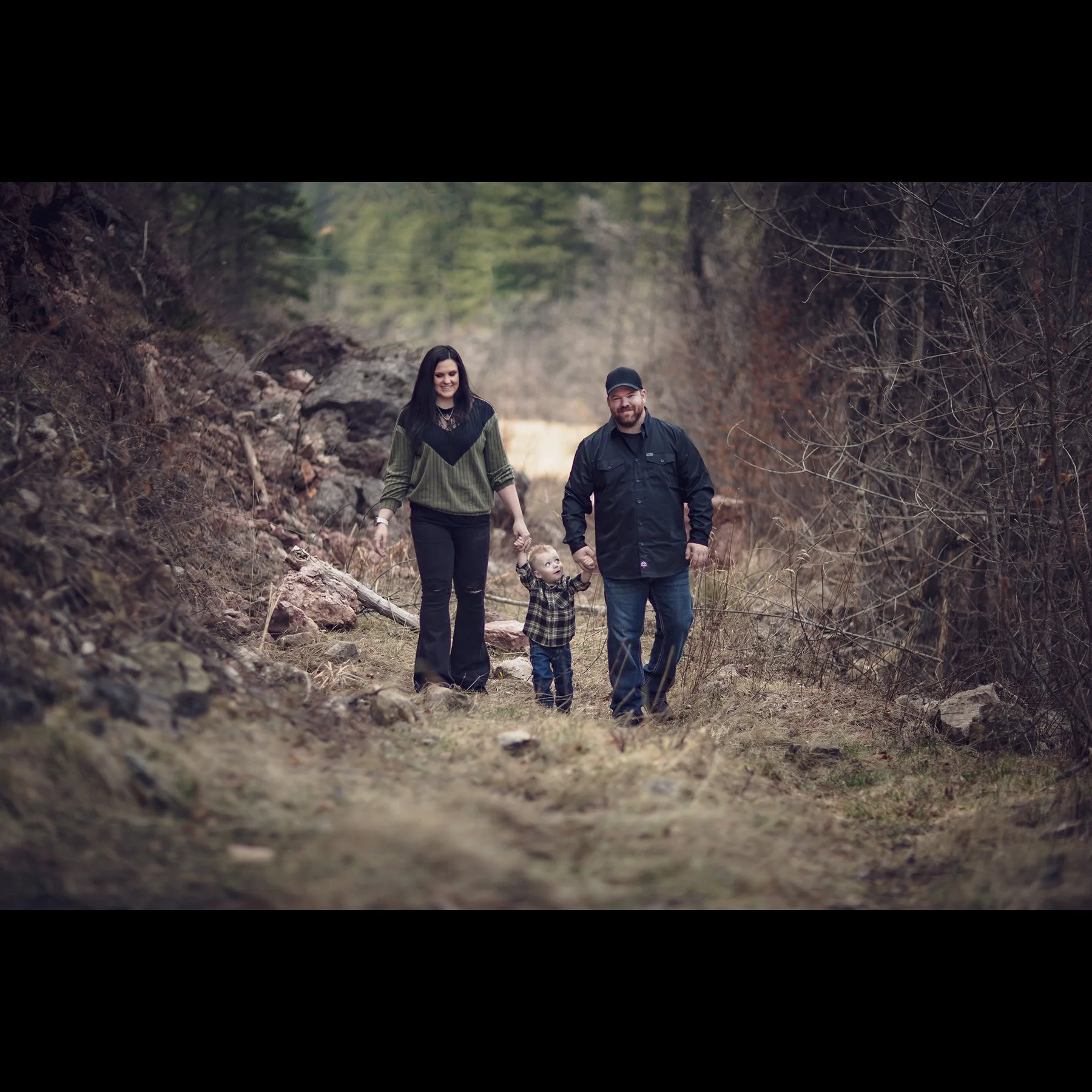 A family of three, a woman, a man, and a small child, walking on a forest trail holding hands, surrounded by trees and rocks.