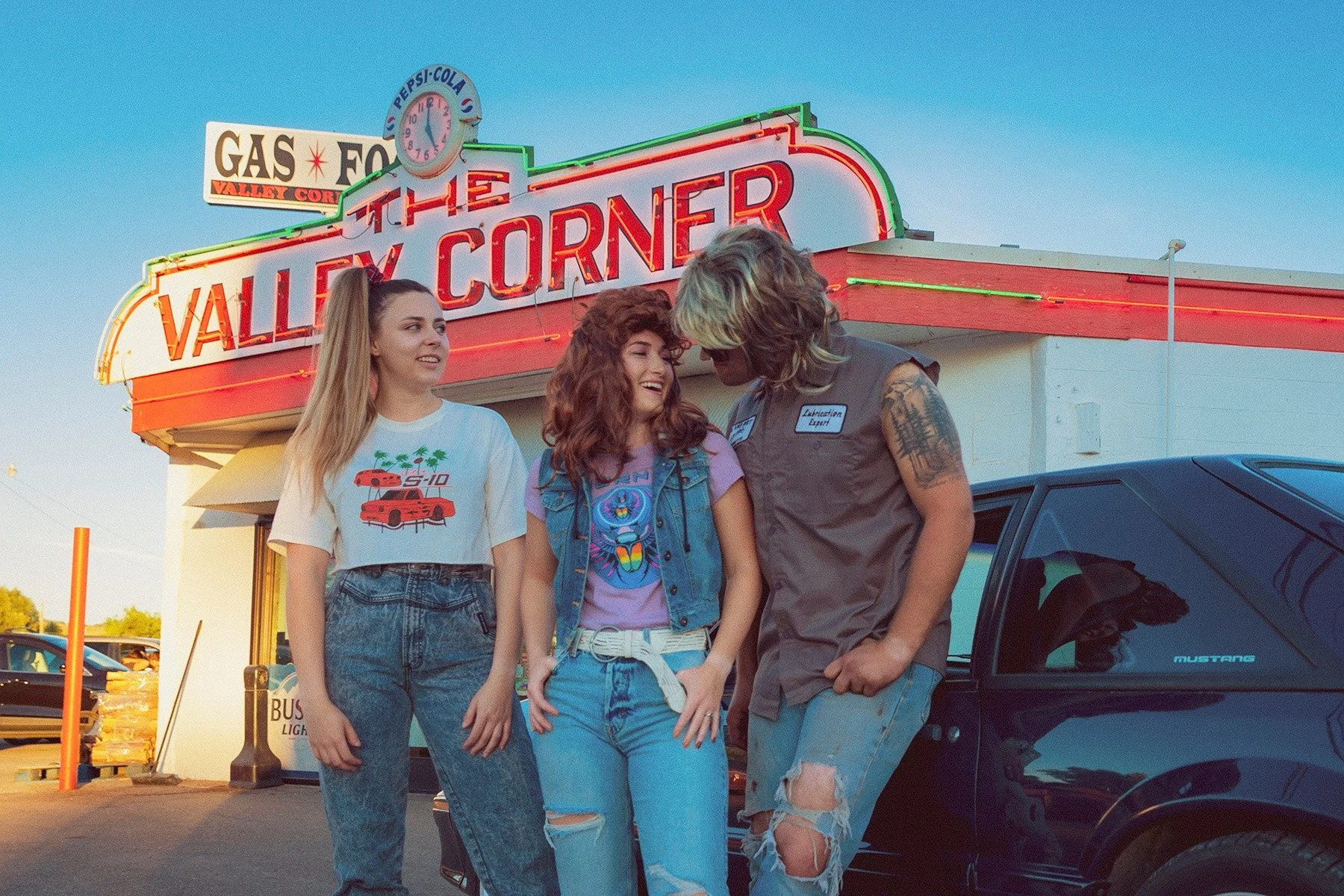 Three young women and a man stand together in front of a neon-lit diner sign reading "Valley Corner" at dusk. The women are smiling and talking, with the man looking down at one of the women. One woman wears a graphic t-shirt and high-waisted jeans, 