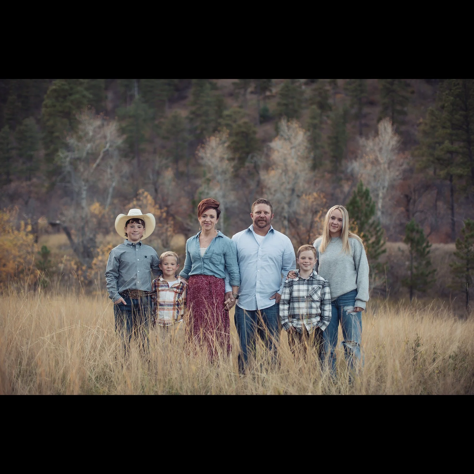 A family of six standing in a field with tall grass, with trees and mountains in the background.