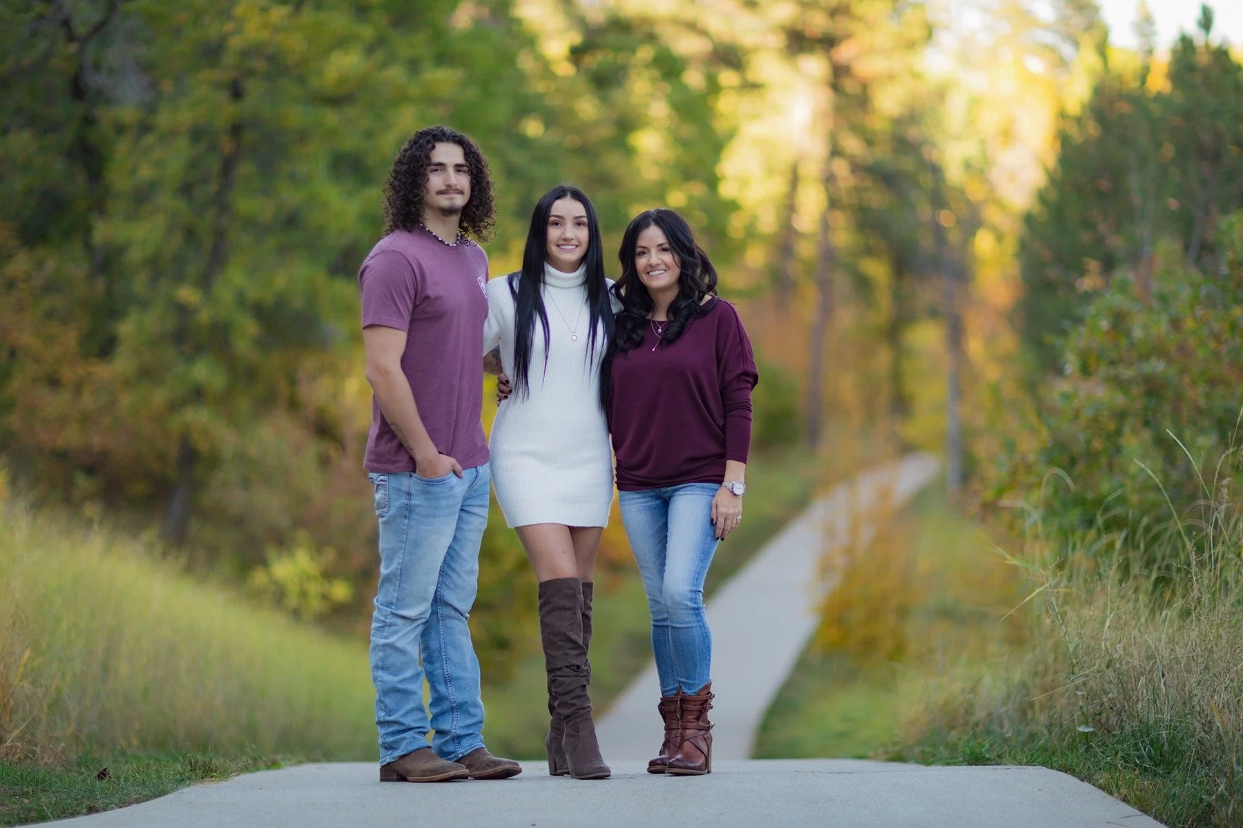 Three people standing on a park path surrounded by trees with fall foliage, smiling at the camera.