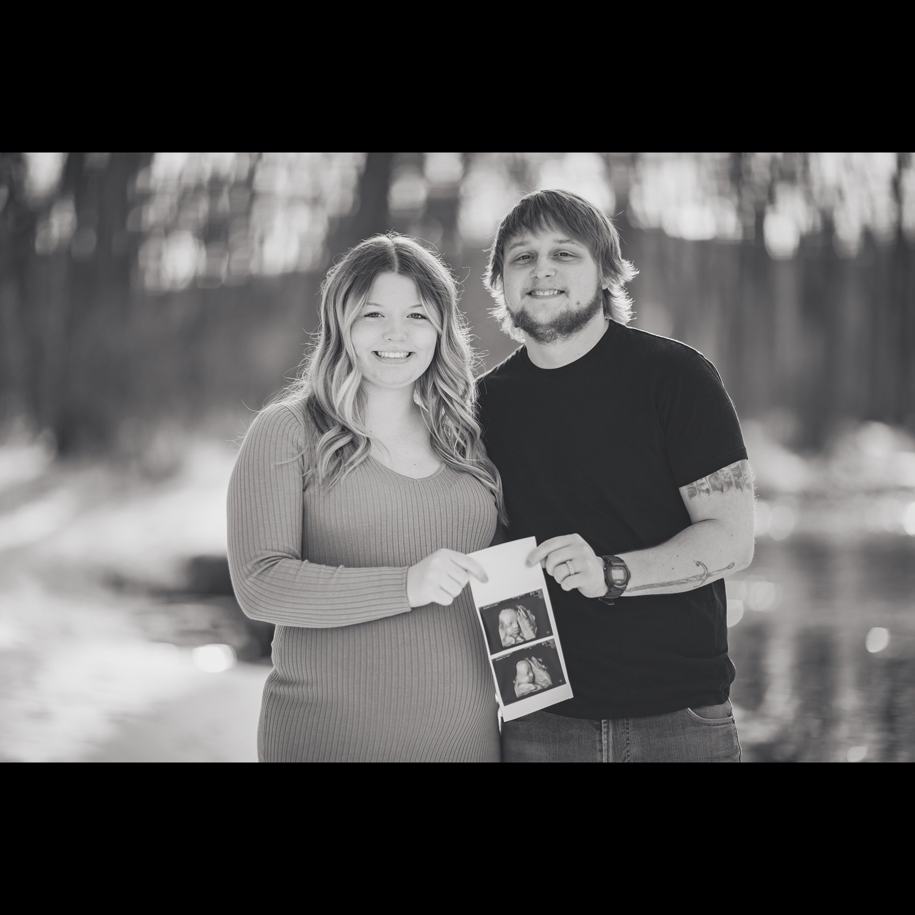 Black and white photo of a pregnant woman and a man smiling while holding ultrasound images outdoors.