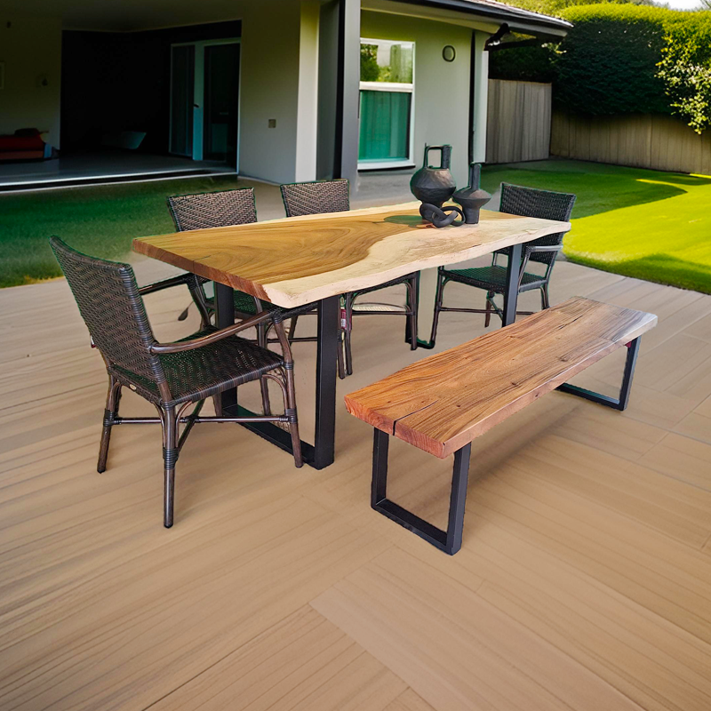 Mesa de madera con cuatro sillas de mimbre y un banco de madera en un patio exterior con césped y una casa moderna al fondo.