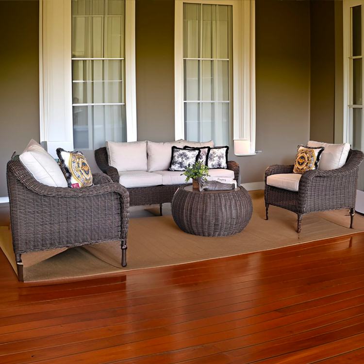 Sala de estar con sillones de mimbre y cojines blancos sobre una alfombra beige, frente a ventanas grandes con cortinas, y piso de madera.