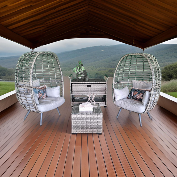 Dos sillones tipo jaula de mimbre con cojines blancos y una almohadilla decorativa, en una terraza con vista a montañas verdes, techo de madera y piso de madera, con mesa central y decoraciones verdes.