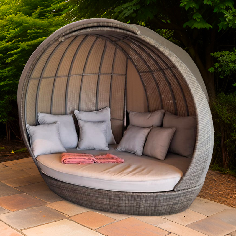 Cabaña de jardín con estructura de mimbre, cojines blancos y una toalla rosa, situada en un entorno natural con árboles verdes.