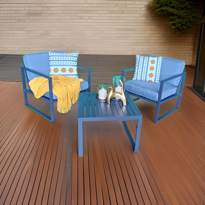 Dos sillas de jardín azul con cojines decorativos en un balcón o terraza con piso de madera, con una mesa azul en medio que tiene decoraciones y un fondo de pared de piedra y plantas.