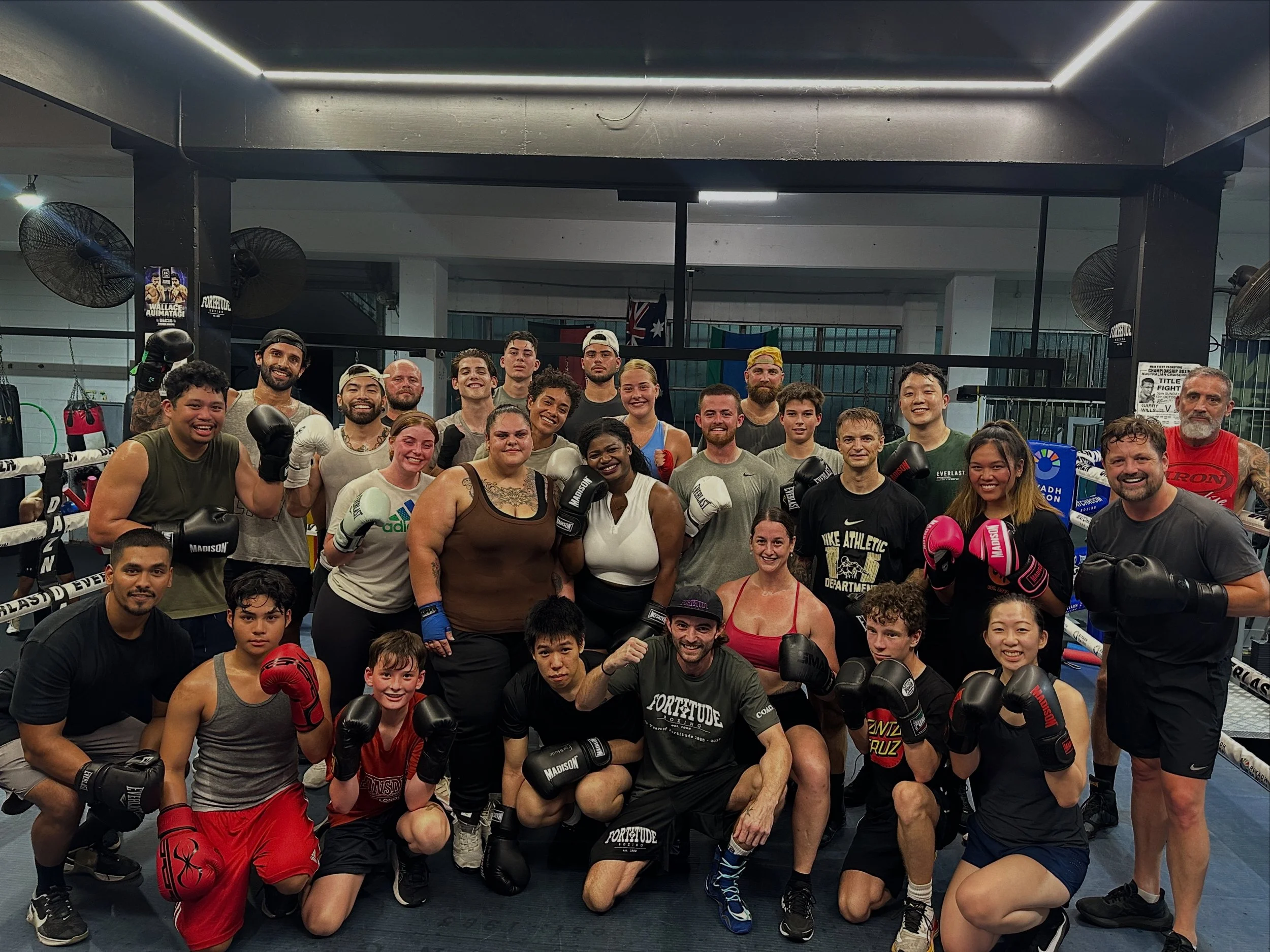 Group of mixed martial artists and trainers in a boxing gym, posing together for a photo. Some are wearing boxing gloves, and there is a boxing ring in the background.
