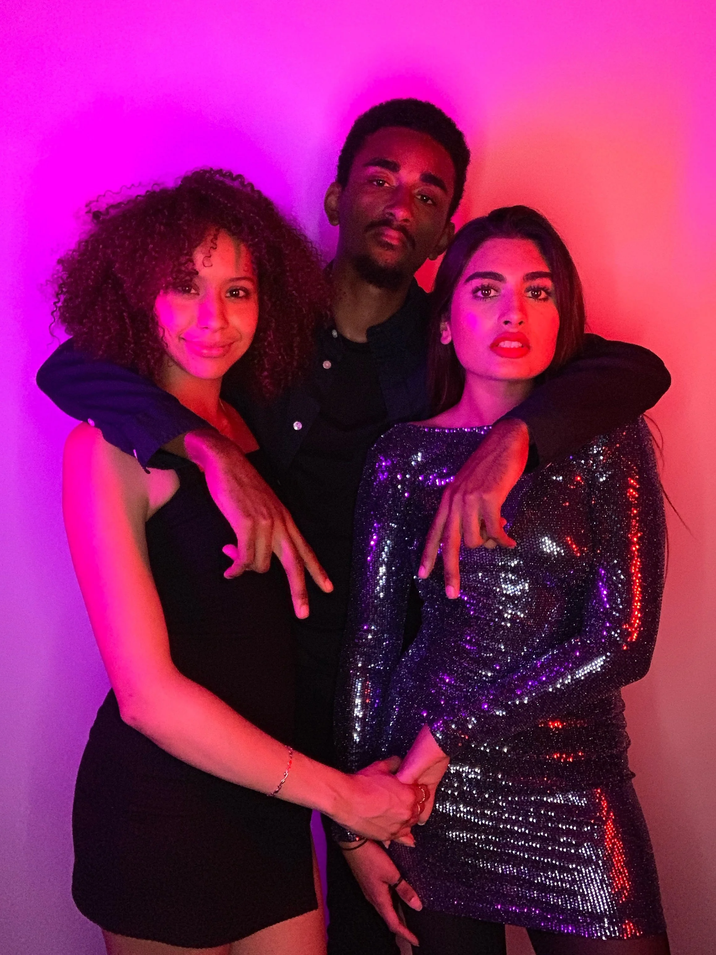 Three people posing together in colorful lighting, with two women and one man standing behind them. The women are touching hands at the bottom, and the man has his arms around the women. The woman on the left has curly hair and is wearing a black dress, the man in the middle has short dark hair and a beard, and the woman on the right has straight hair and is wearing a sparkly dress.
