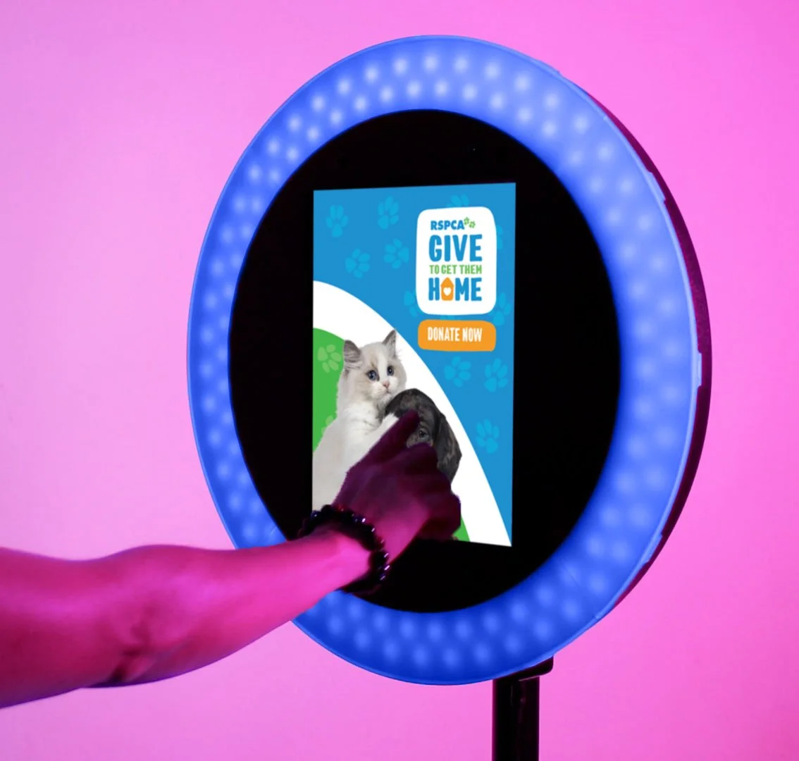 Person touching a screen promoting an animal shelter with a blue ring light around it, featuring a cat and a dog, and a message encouraging donations.