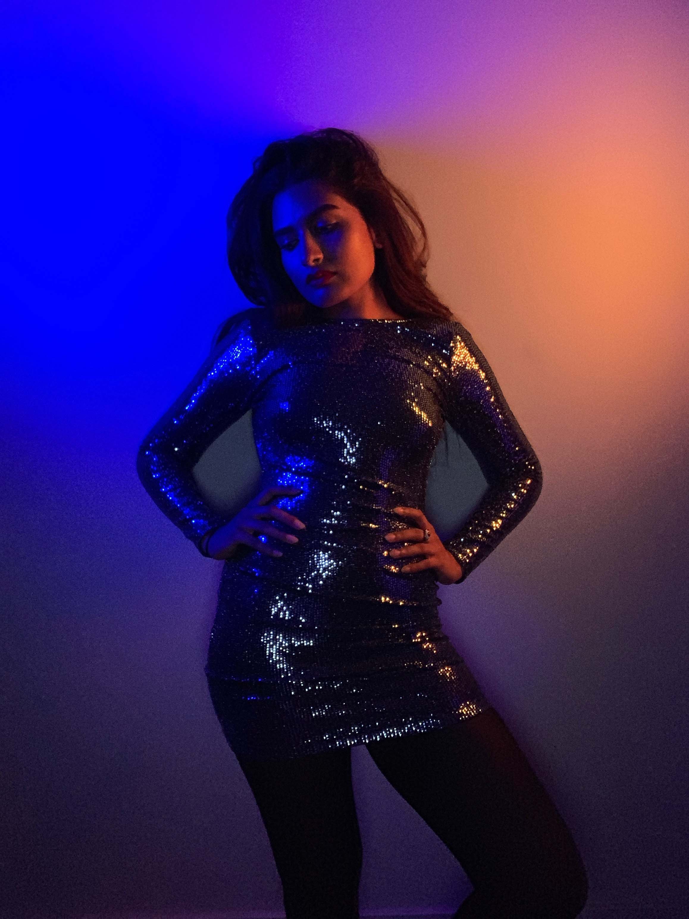 Woman in a shiny, silver sequined dress standing with hands on hips, against a multi-colored, gradient background.