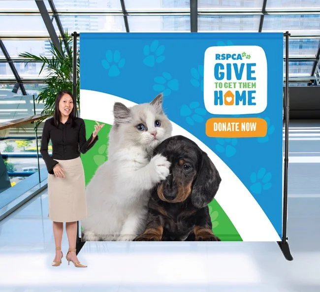 Premium Ausome Love Studio photo booth banner display featuring a kitten and puppy, showcased in a modern glass building for a charity event.