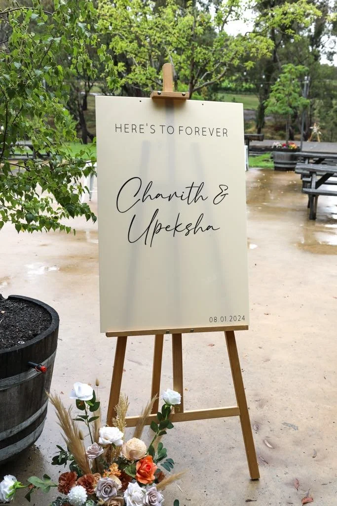 Wedding sign on easel outdoors, reads 'Here’s to Forever' with names and date, surrounded by flowers and greenery.