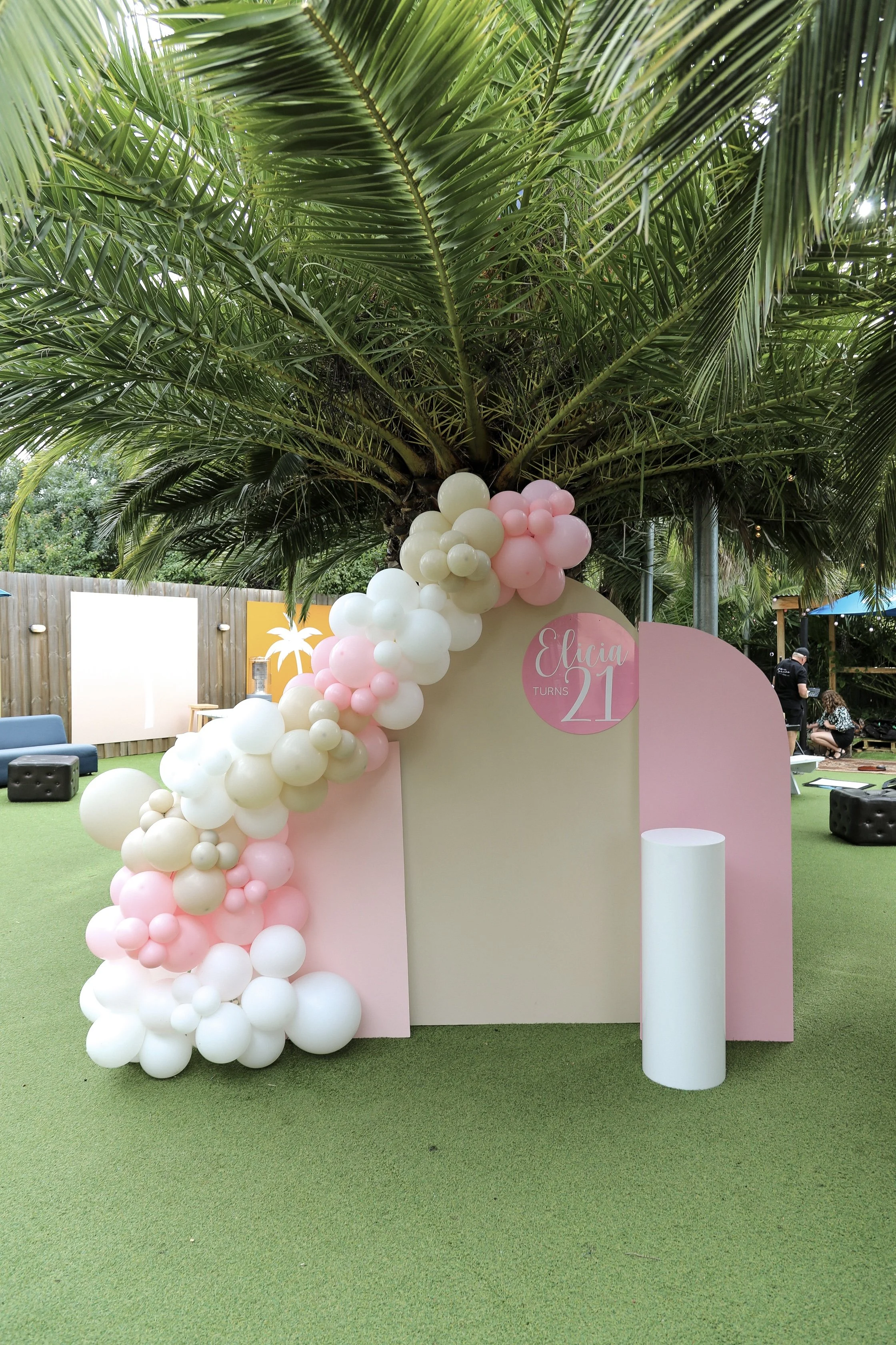 Outdoor birthday party setup with balloon decorations, a palm tree, and a sign reading "21."
