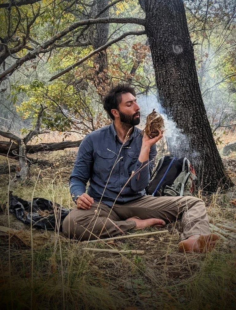 A man sitting cross-legged on the ground in a forested area, holding a burning bundle of leaves or herbs near his face, with smoke rising from it.