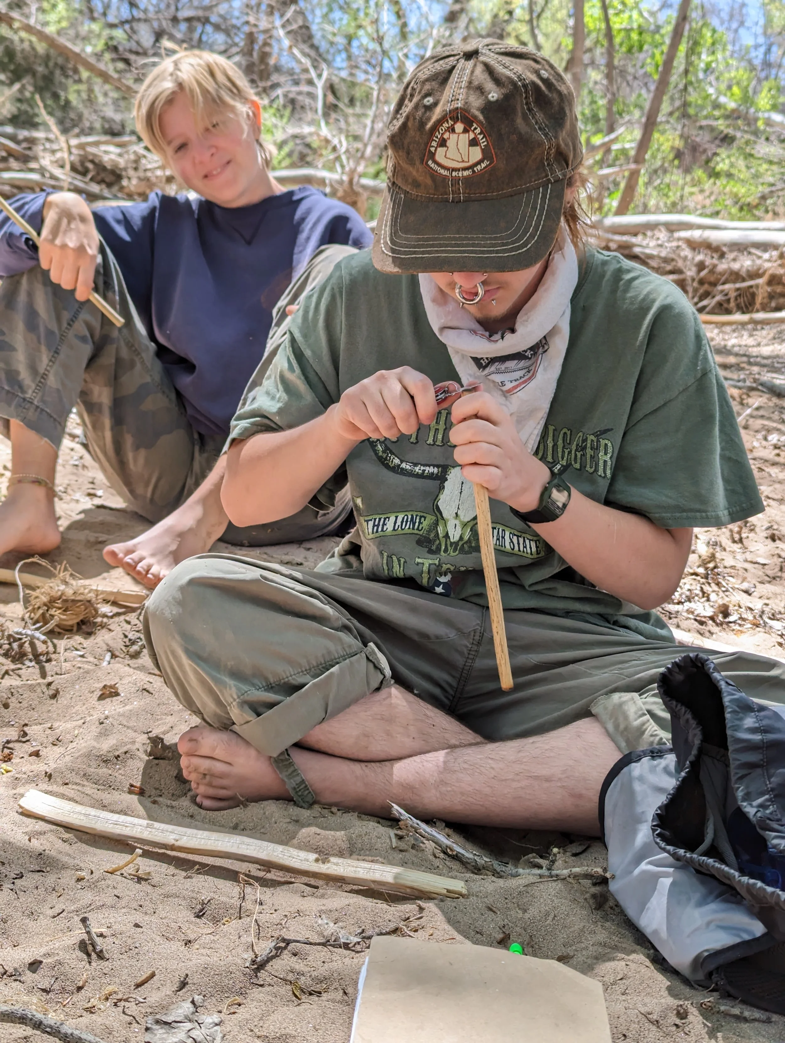 Two young men with piercings sitting in the sand outdoors. One is wearing a green t-shirt and a cap, handling a small tool, with a backpack nearby. The other is in a dark sweater with camo pants, sitting behind and smiling at the camera. They are in a wooded area.