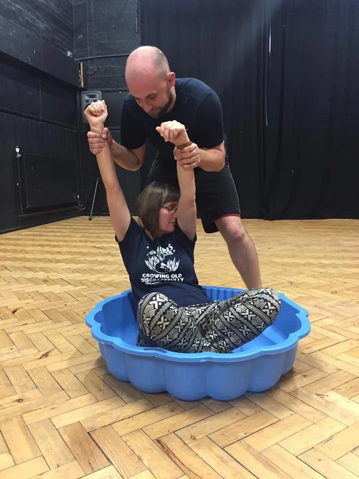 A person sitting in a blue plastic tub on a wooden floor, with another person standing behind them holding their arms up. The sitting person is wearing a black shirt and patterned pants.