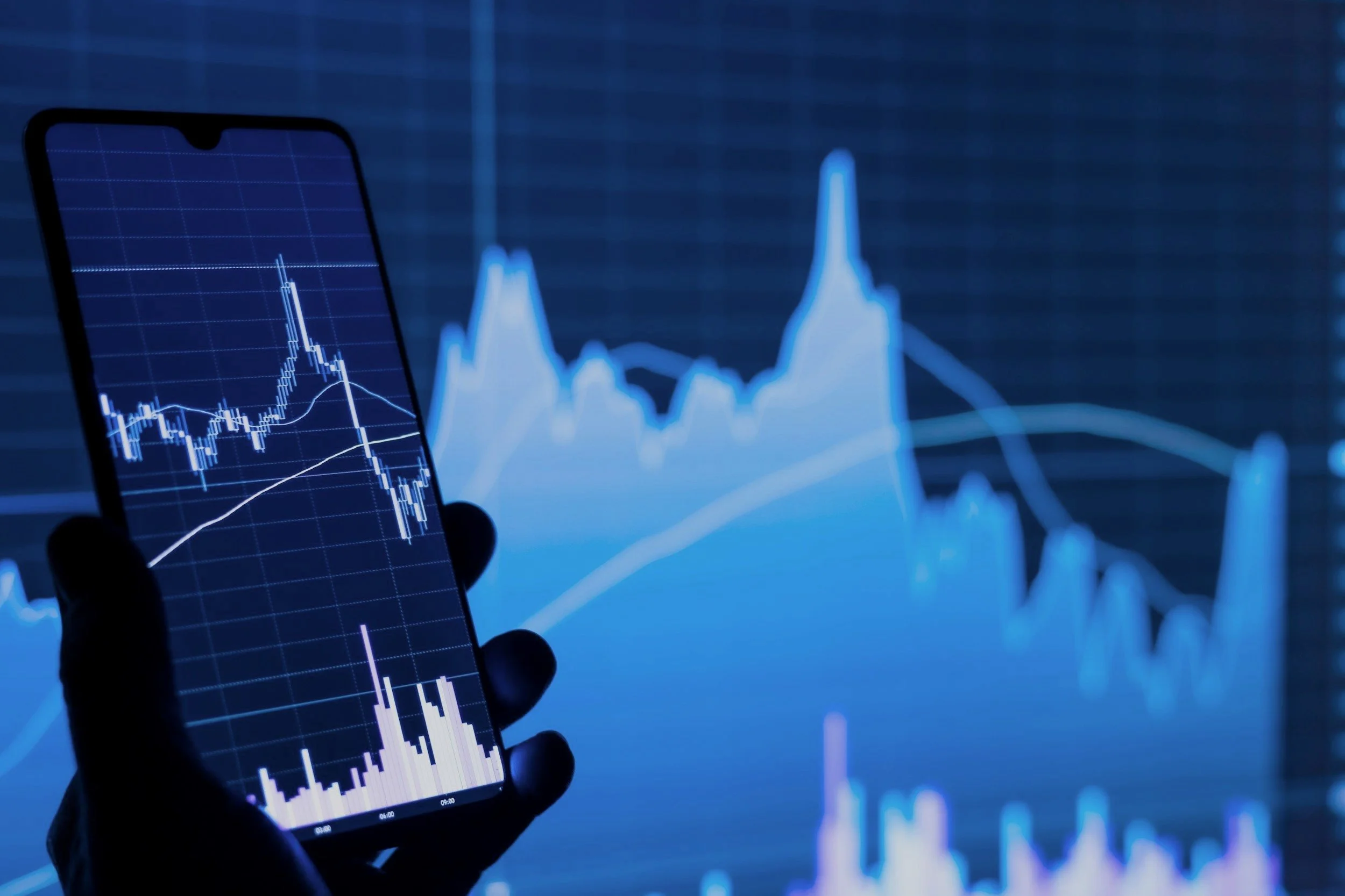 A hand holding a smartphone displaying a line graph with candlestick patterns, with a large screen in the background showing a financial chart with peaks and valleys, indicating stock or cryptocurrency market data.