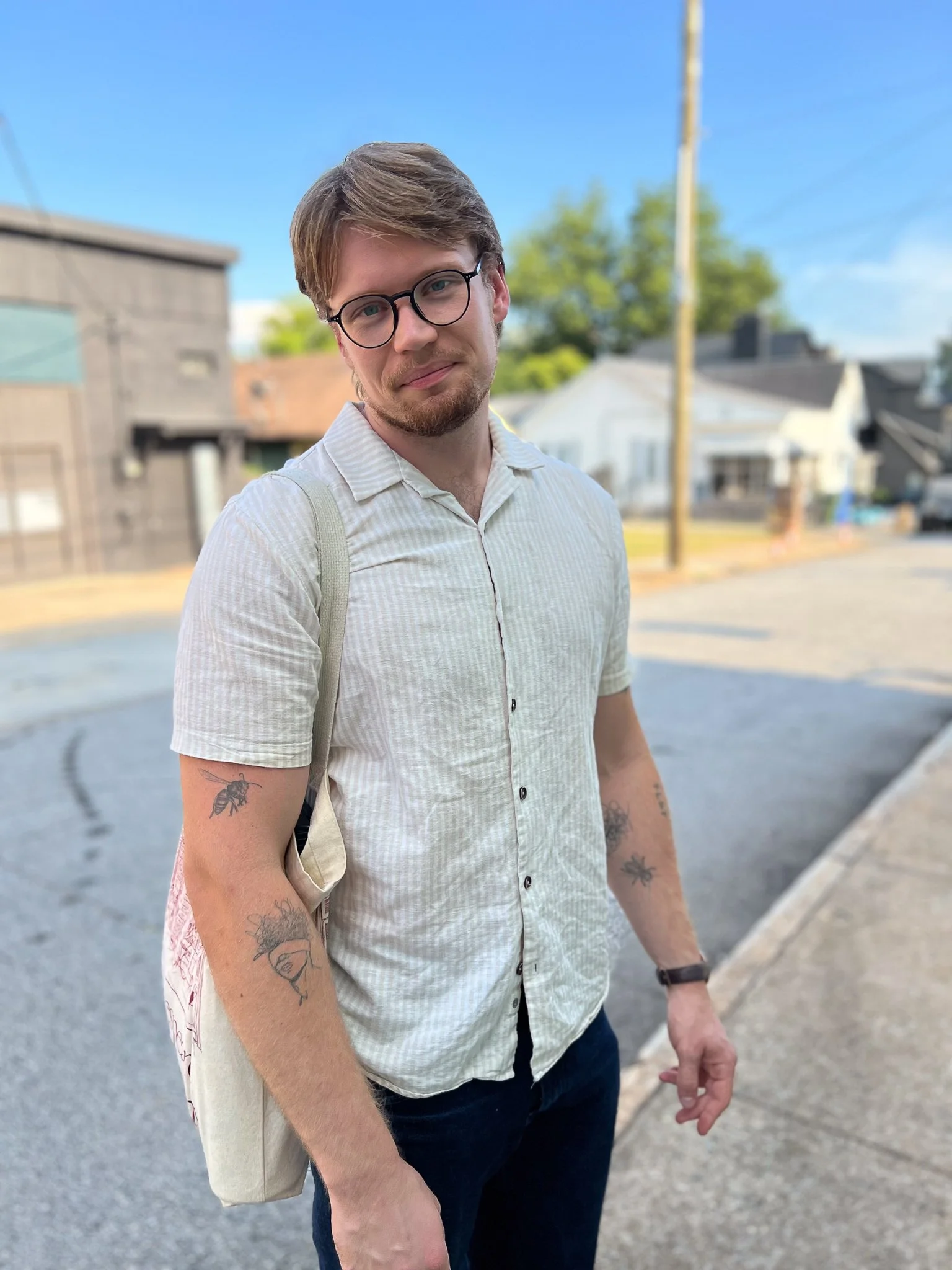 Young man with glasses and tattoos on arms standing outdoors on a sunny day, wearing a light-colored short-sleeve shirt and backpack, in a neighborhood with houses and a blue sky in the background.