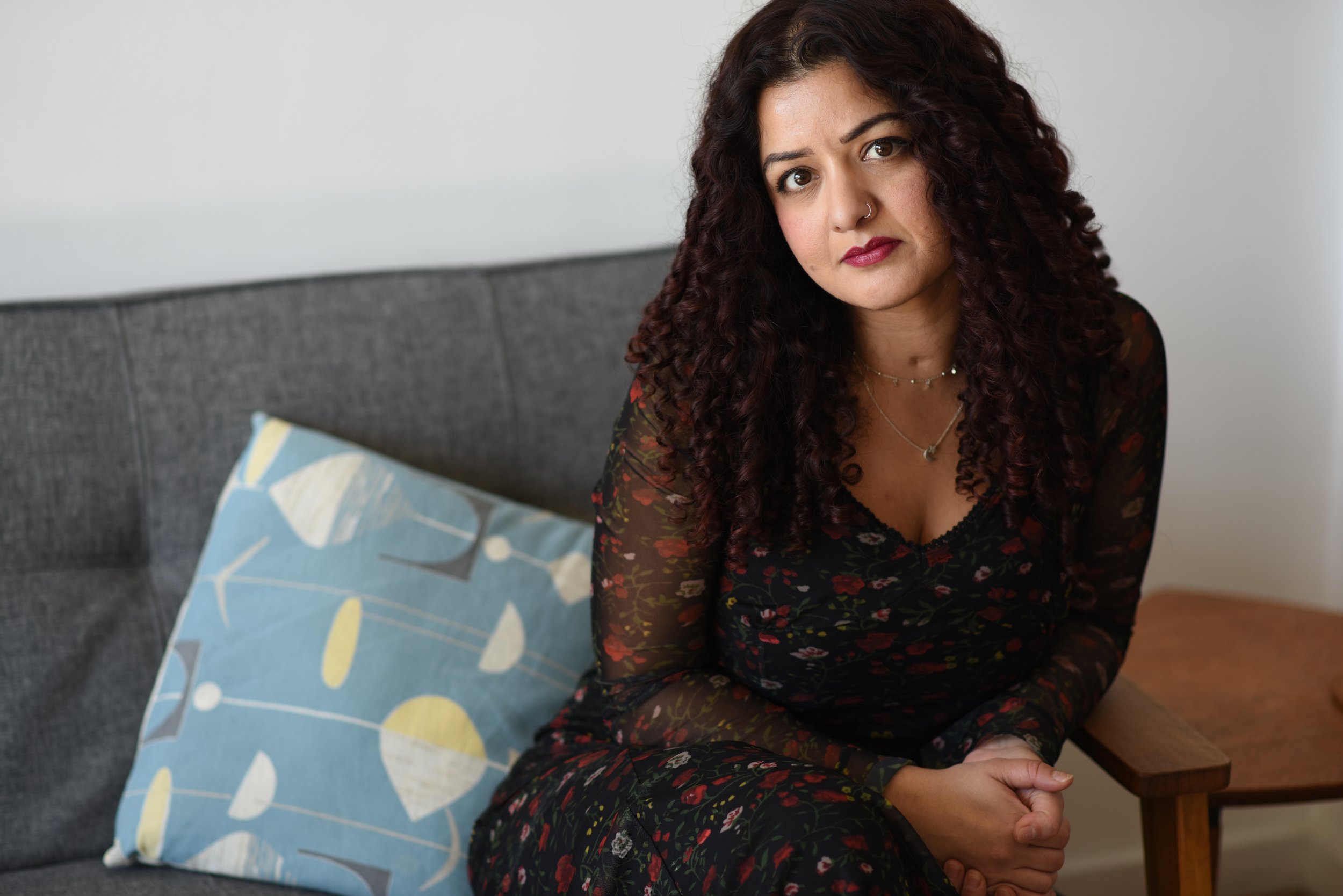 A woman with curly dark brown hair and a nose ring sitting on a gray couch, wearing a black floral dress, with a blue-patterned pillow beside her in a cozy living room.