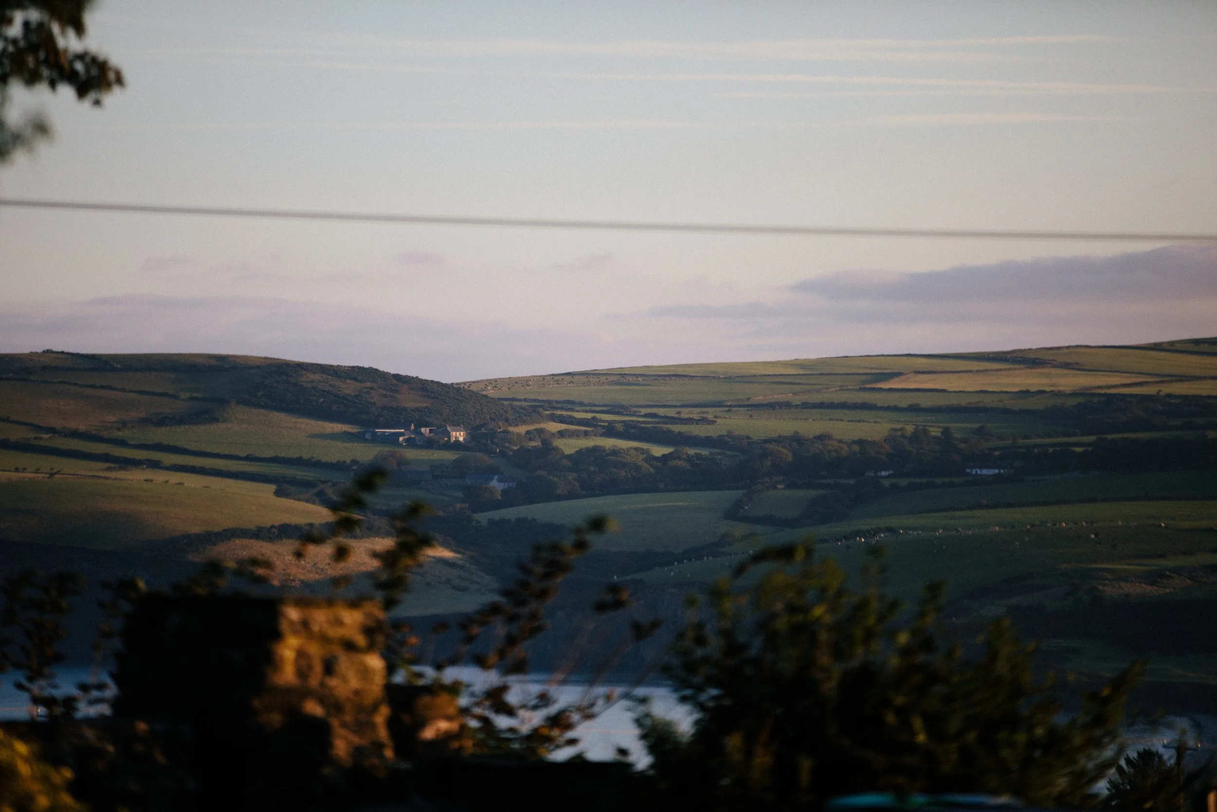A scenic landscape showing rolling green hills with a few houses in the distance, with a partly cloudy sky overhead during daytime.