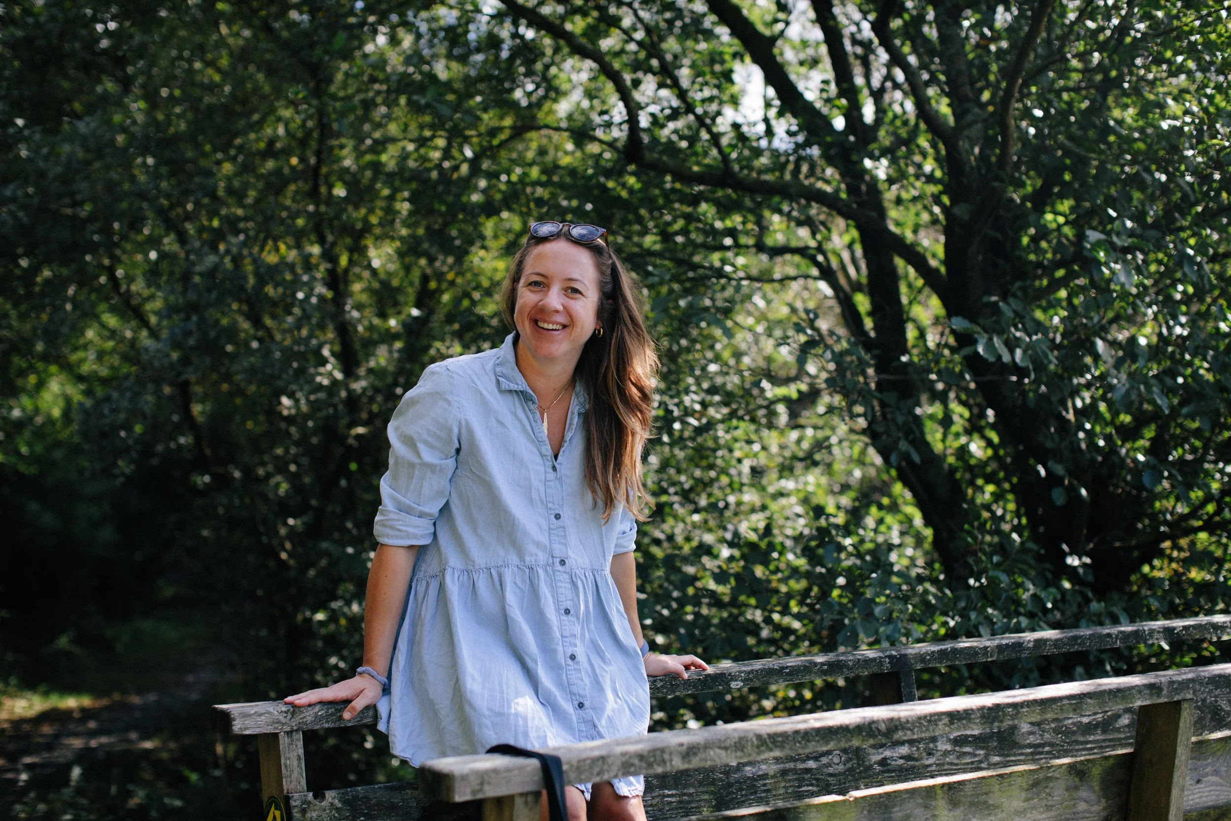 A woman with long brown hair, wearing a light blue dress and sunglasses on her head, leaning on a wooden fence in a lush green park, smiling at the camera.