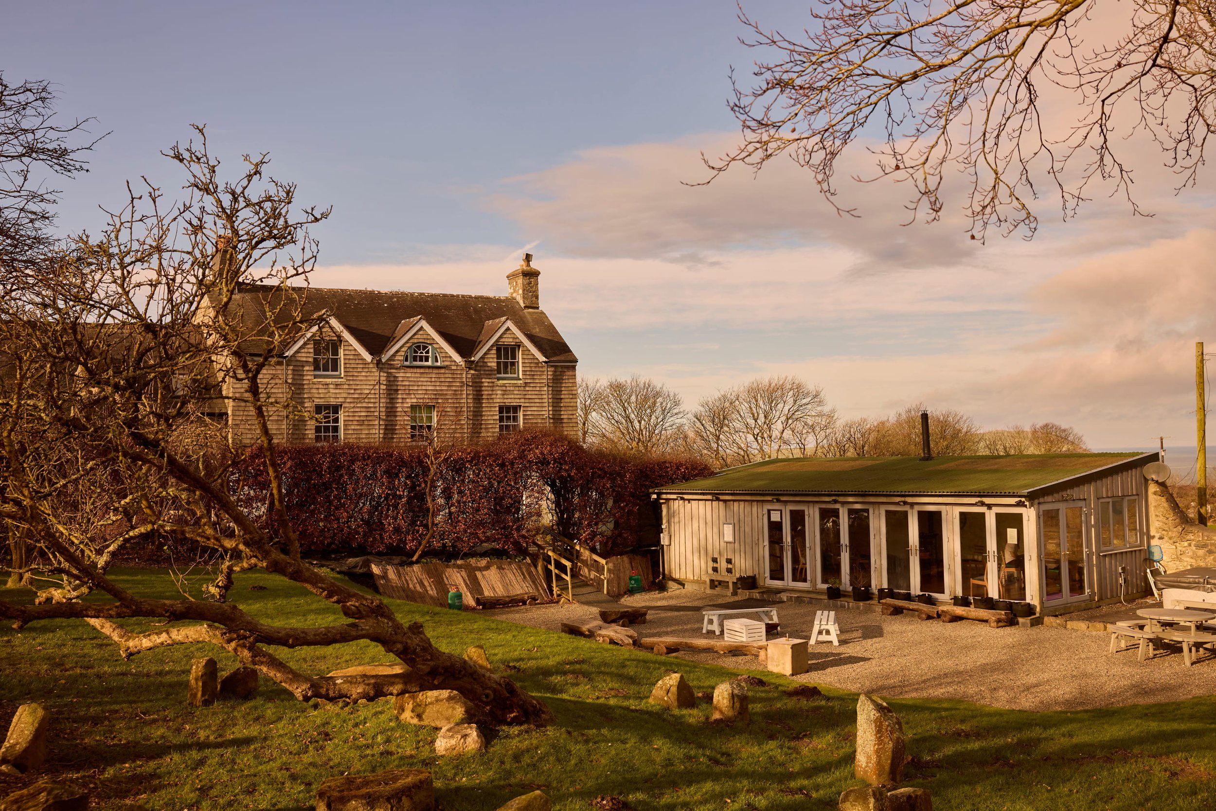 A house with wood shingles and a chimney surrounded by leafless trees and a grassy lawn. There is a small building with windows and a door in the foreground, and outdoor furniture such as benches and chairs. The sky is partly cloudy with hints of sunlight.