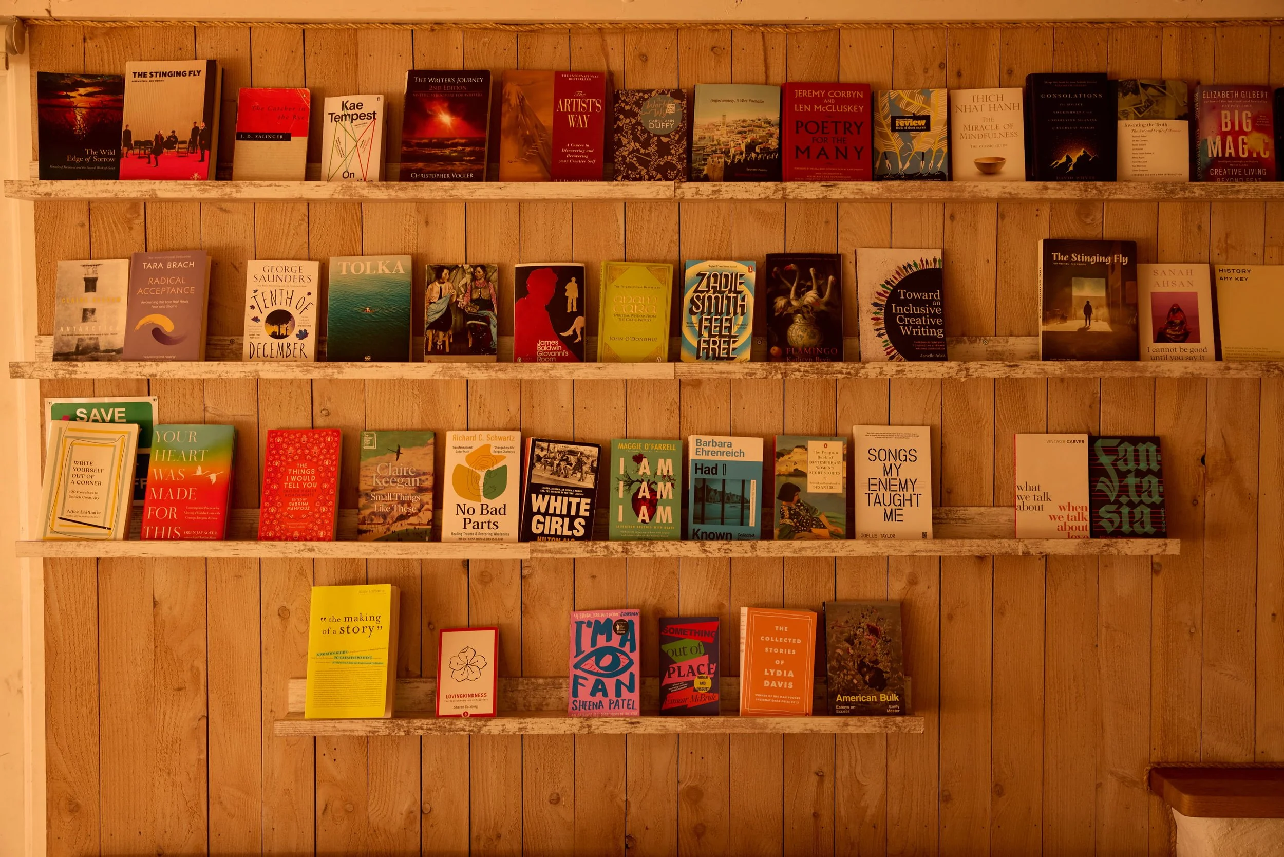 Multiple books arranged on three wooden shelves against a wooden wall. The top shelf contains mostly hardcover books, and the lower shelf has a mix of hardcover and paperback books with larger titles visible, including 'Your Heart Was Made for This' and 'No Bad Parts'.