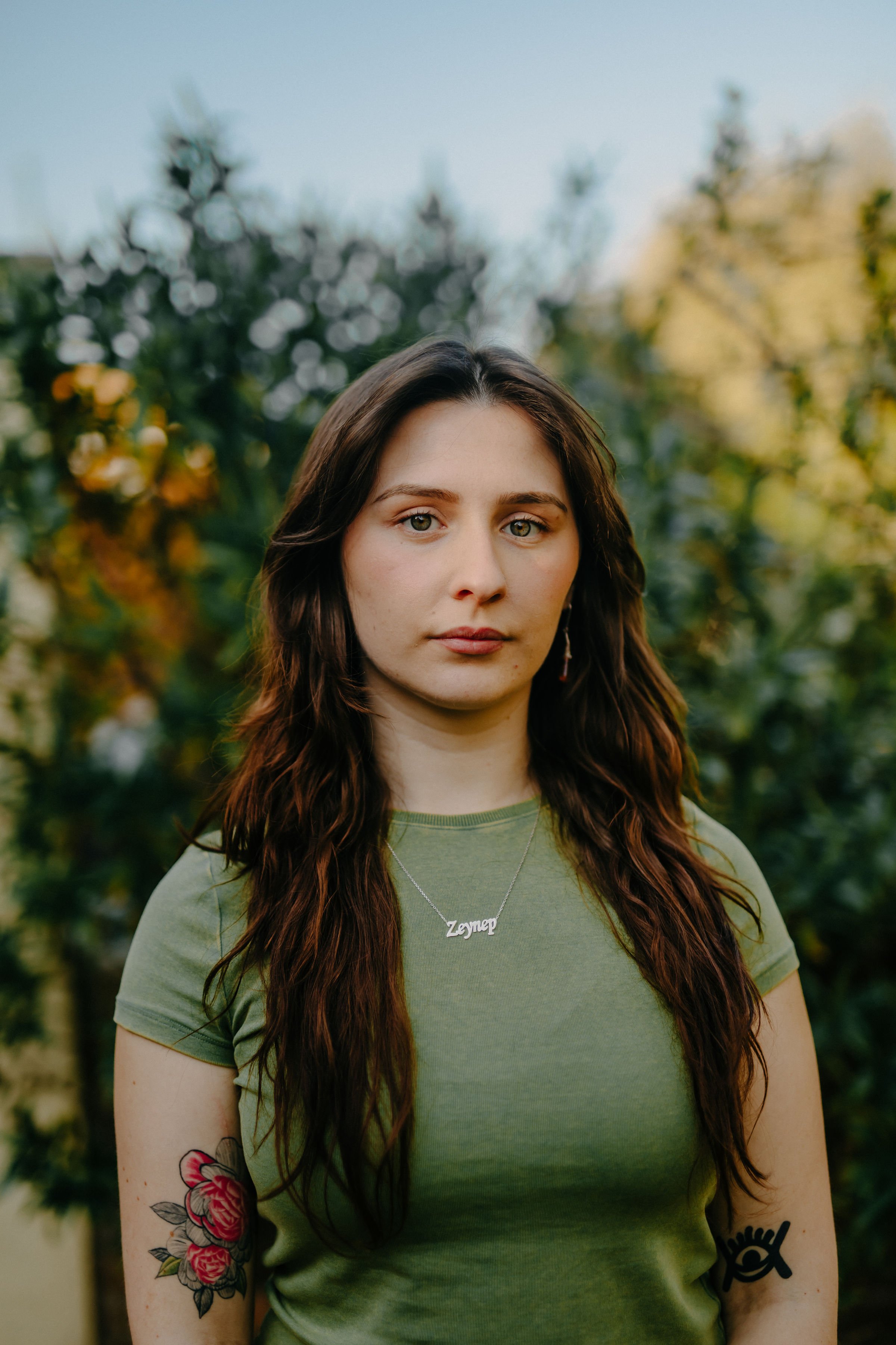 A young woman with long brown hair and fair skin stands outdoors in front of green foliage, wearing a green t-shirt and a necklace with the name "Zeynep." She has tattoos on her left arm, including a bouquet of flowers and a black eye design. The background is slightly blurred, featuring trees and a clear sky.