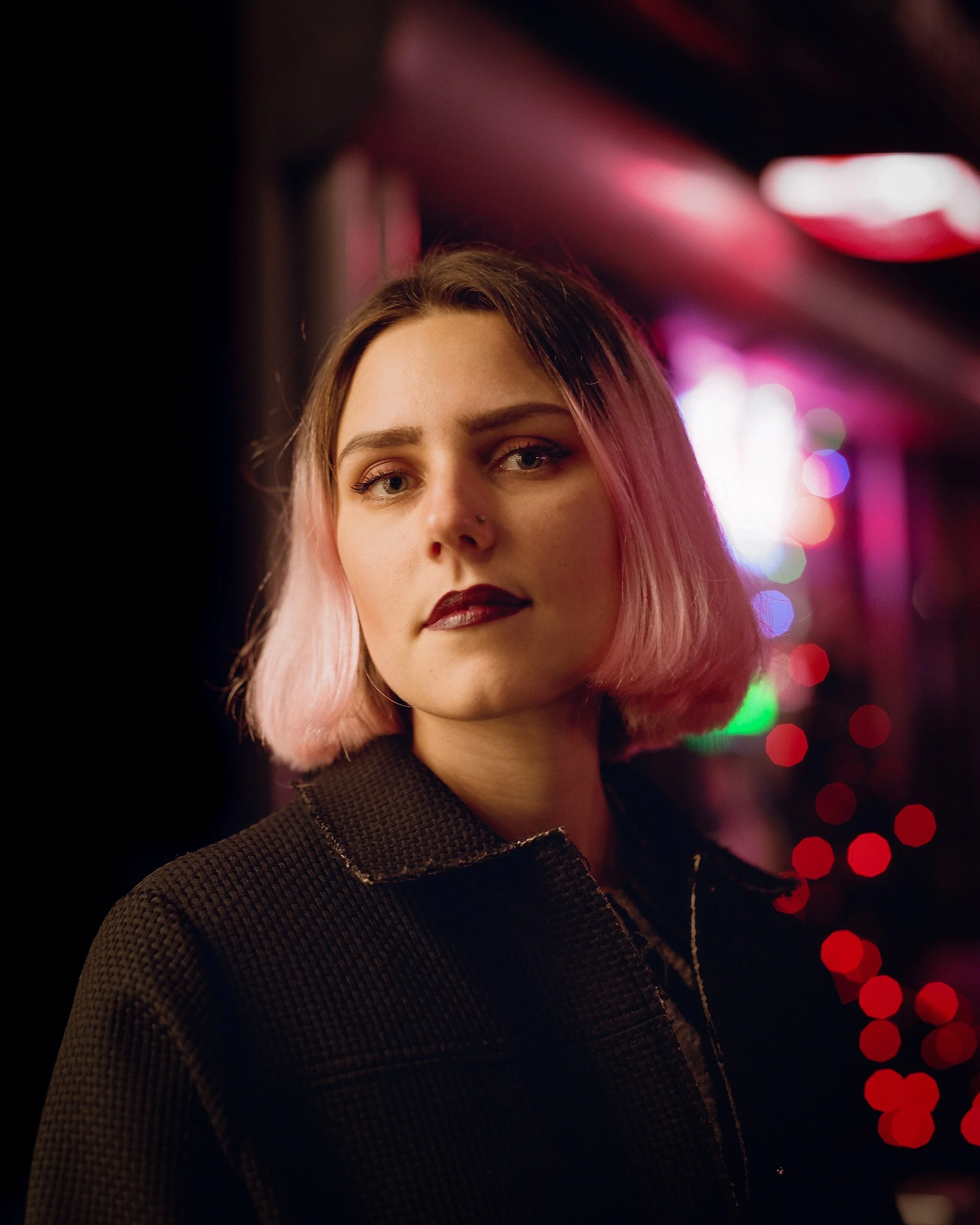 A young woman with pink and brown hair, wearing dark lipstick and a black textured coat, standing in front of a background with colorful bokeh lights at night.