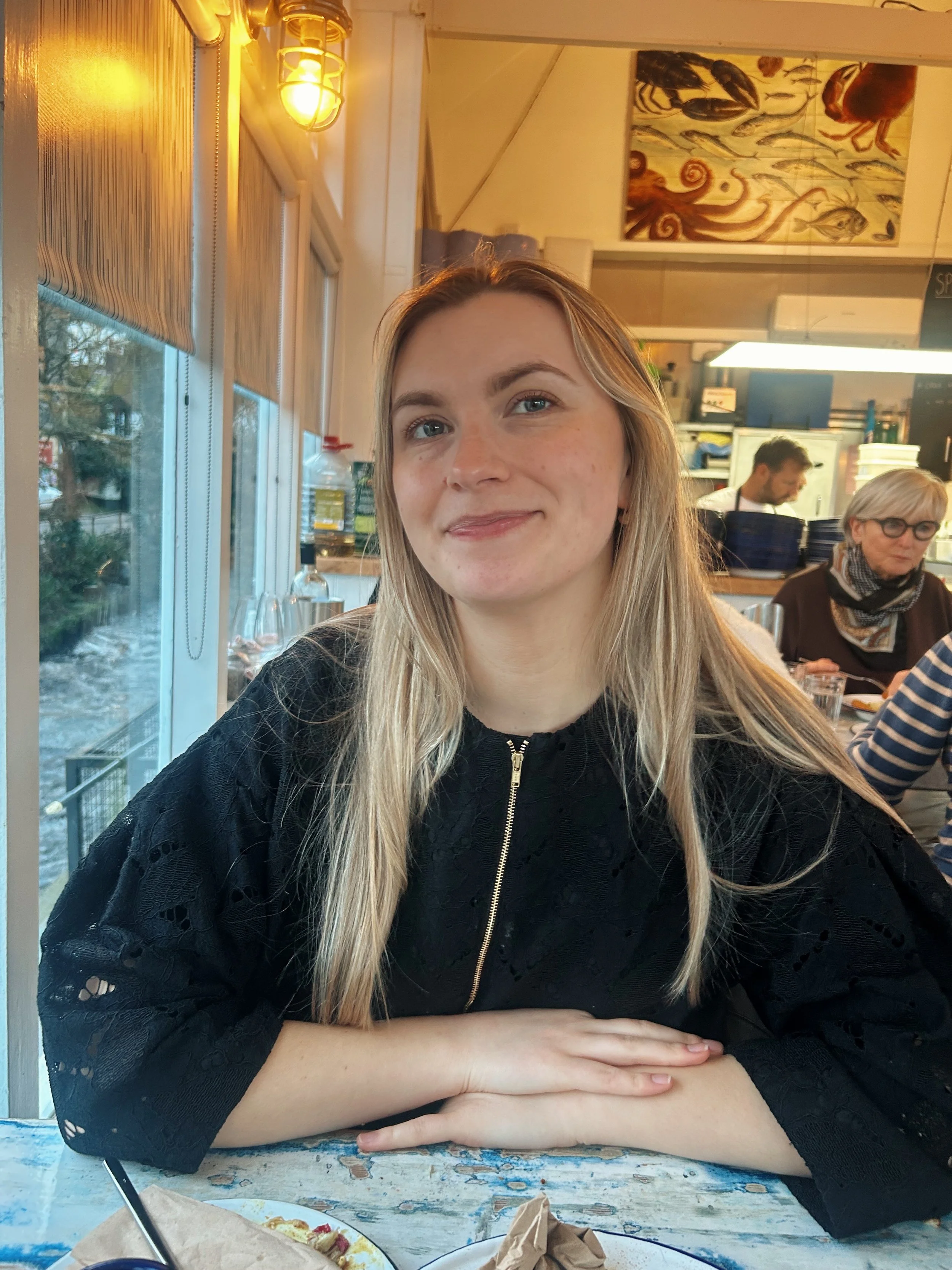 A young woman with long blonde hair and light skin sitting at a rustic restaurant table, smiling slightly. Behind her, a window shows trees outside. To her right, an older woman with glasses and a scarf sits at the counter, focused on eating. The restaurant has warm lighting, art on the wall, and a busy, cozy atmosphere.