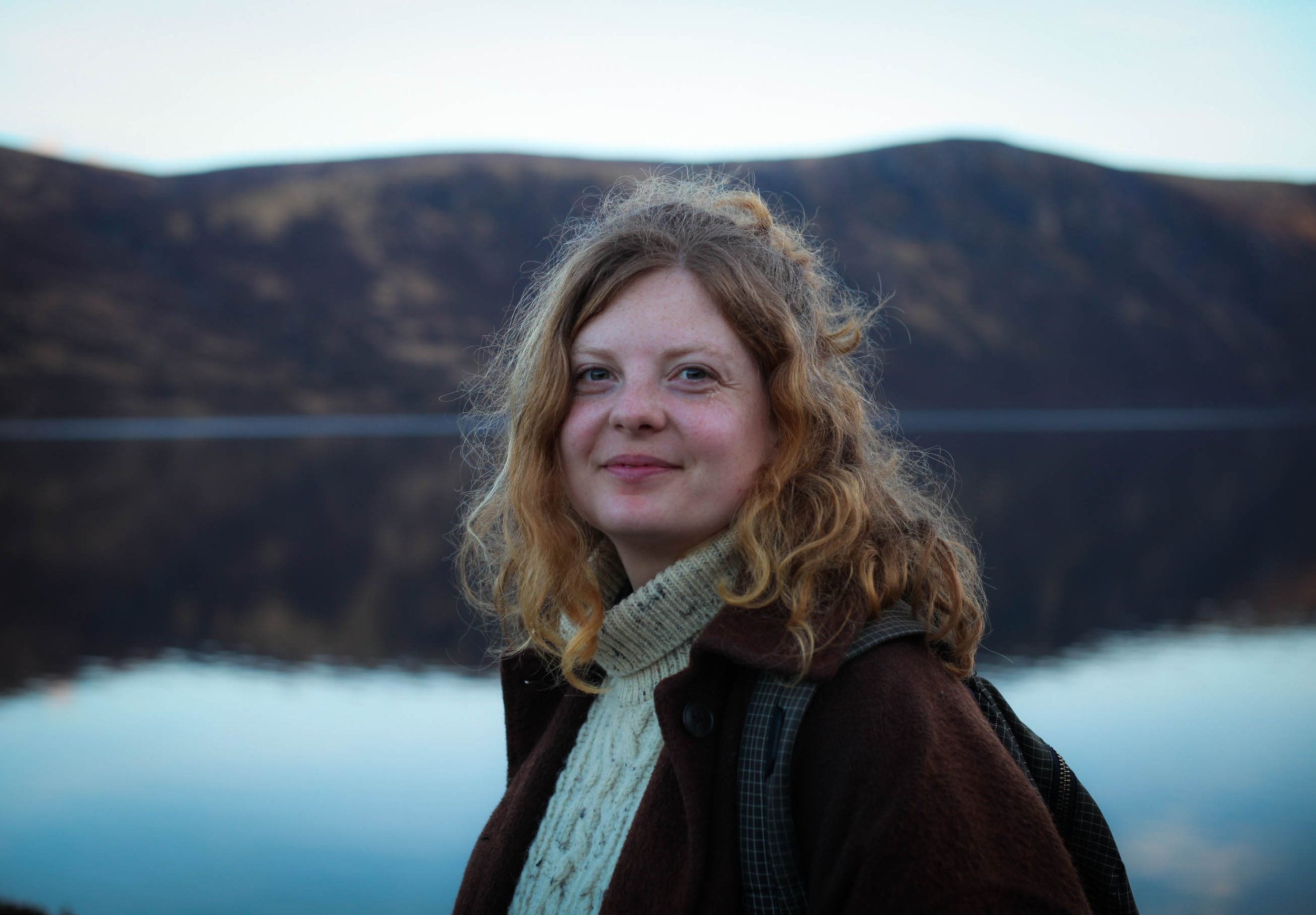 A woman with curly red hair and fair skin standing outdoors near water, with mountains in the background during daytime.