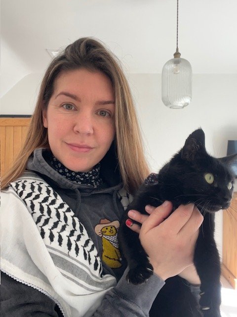 A young woman holding a black cat indoors with a pendant light hanging from the ceiling.