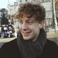 Young man with curly hair smiling, standing outdoors with historic buildings and crowd in the background.