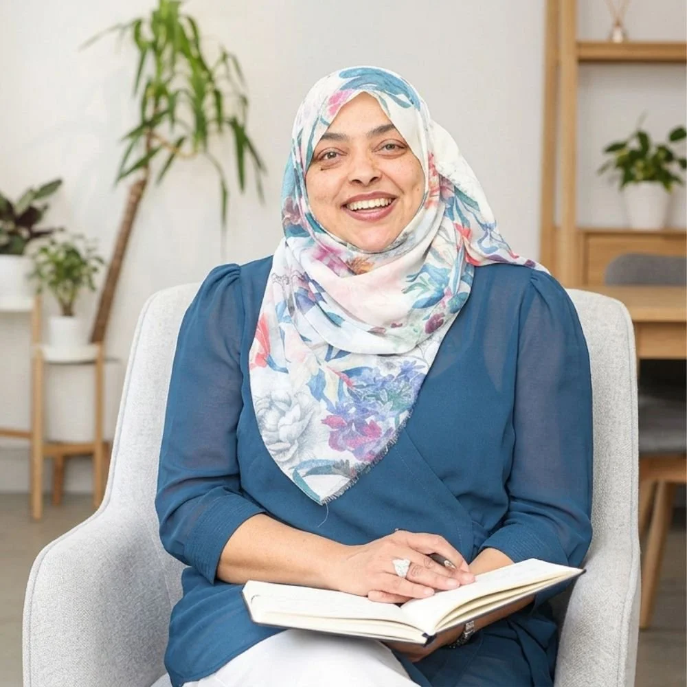 Nurjahan Khatun, coach & speaker. smiling in a floral hijab and blue shirt sitting in a chair with an open notebook on her lap in a room with houseplants and wooden furniture.