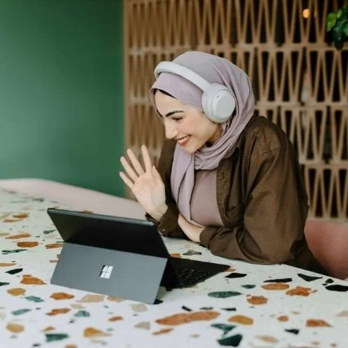Nurjahan Khatun, coach & speaker. A woman wearing a hijab and headphones smiling at a tablet on a table.