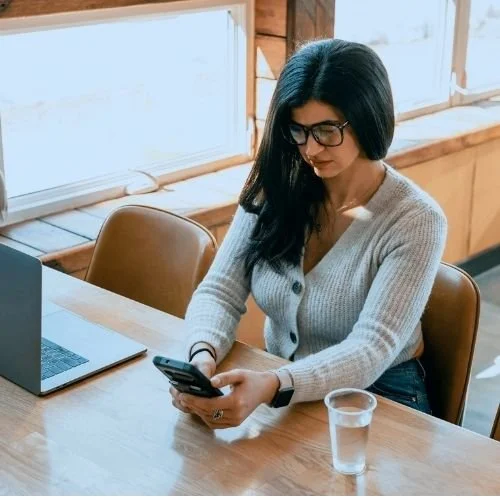 Woman with black hair and glasses sitting at a wooden table, looking at her phone with a glass of water nearby, inside a bright, wood-paneled room. Nurjahan Khatun, coach & speaker.