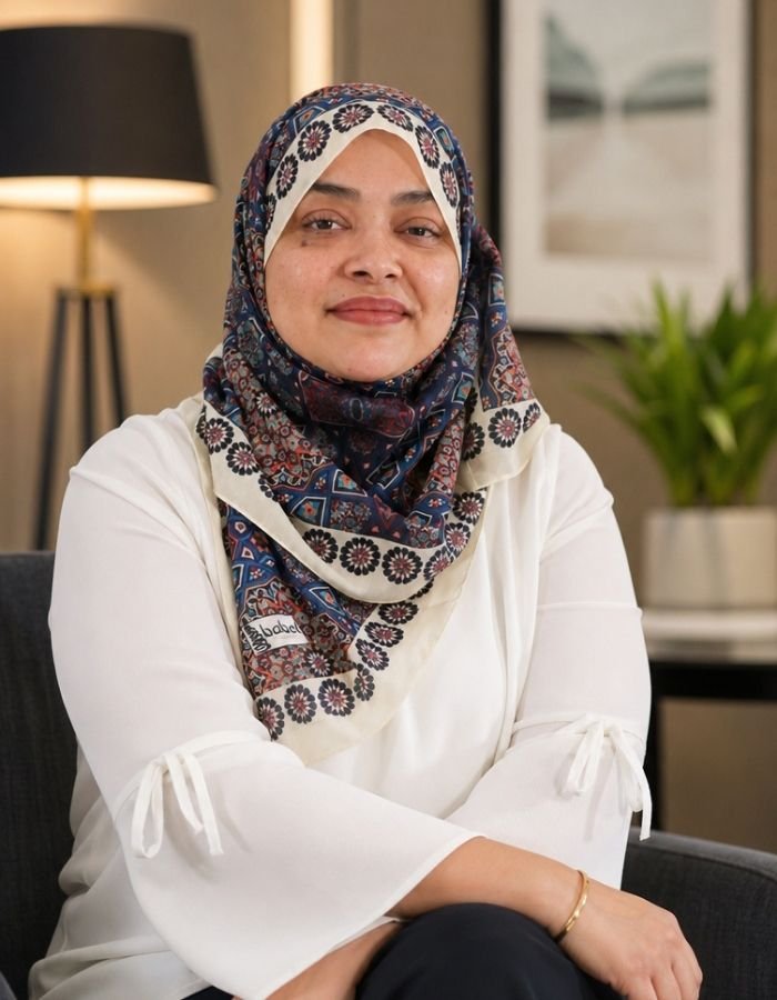 Nurjahan Khatun, coach & speaker. wearing a patterned hijab and white blouse sitting on a chair in a living room with a lamp and a plant in the background.