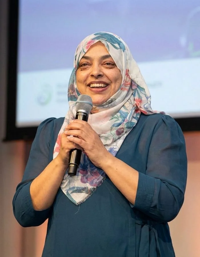 Nurjahan Khatun, coach & speaker wearing a floral hijab is smiling and holding a microphone during a speech or presentation.