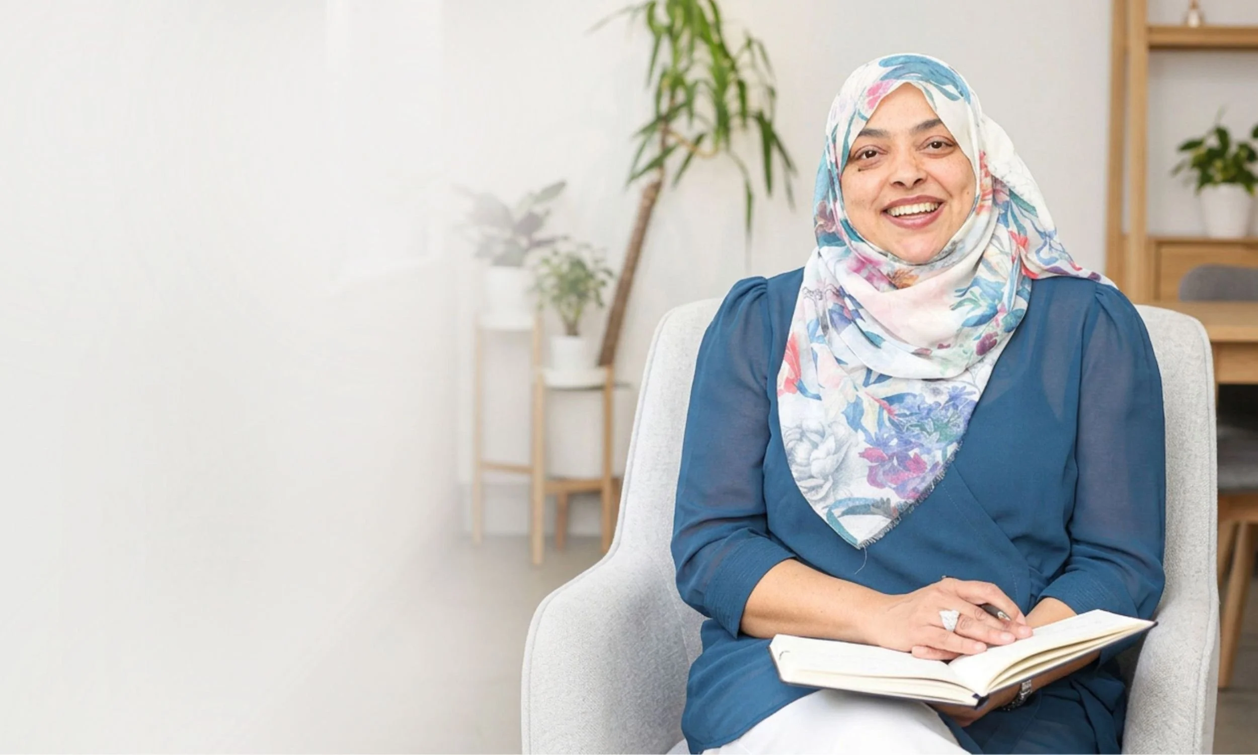 Nurjahan Khatun, coach & speaker  wearing a floral hijab and blue top smiling while sitting in an office chair, holding an open book. The background shows a white wall with a wooden shelf and potted plants.