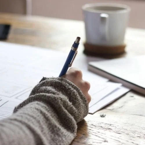 A person wearing a gray knitted sleeve holding a blue pen and writing on papers on a wooden desk, with a white mug and notebook nearby. Nurjahan Khatun, coach & speaker.