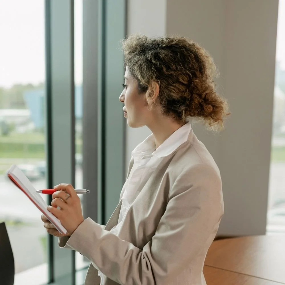 Woman with curly hair and a beige blazer standing near window, writing in a notepad with a red pen. Nurjahan Khatun, coach & speaker.