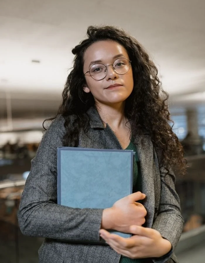 A young woman with curly dark hair and glasses, wearing a gray blazer, holding a blue folder and looking at the camera in an indoor setting. Nurjahan Khatun, coach & speaker.