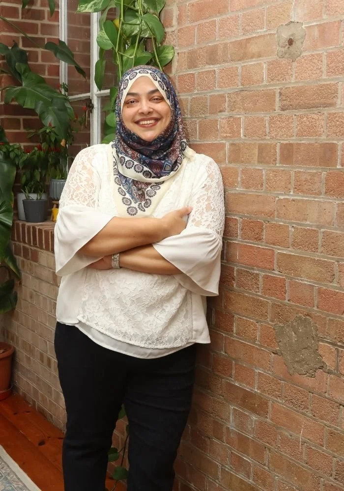 Nurjahan Khatun, coach & speaker. in a white lace top and black pants, wearing a patterned hijab, smiling and standing against a brick wall with green plants in pots behind her.