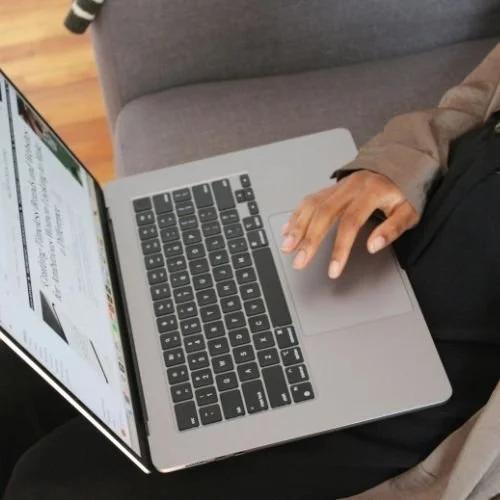 Person sitting on a gray couch using a laptop on their lap, with fingers on the keyboard and a webpage open on the screen. Nurjahan Khatun, coach & speaker.