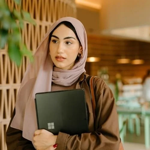 Young woman with a hijab holding a black tablet in a modern office building. Nurjahan Khatun, coach & speaker.