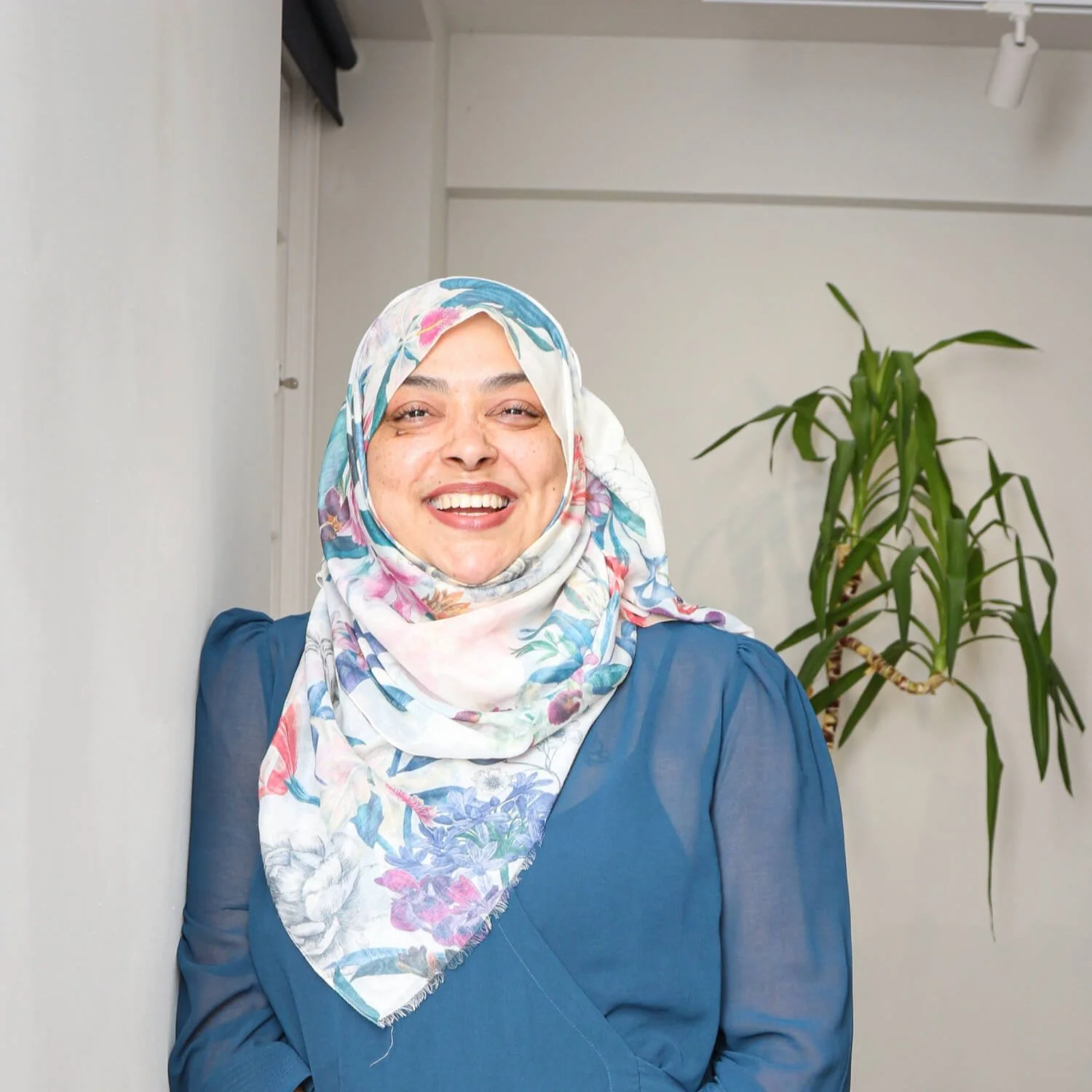 Nurjahan Khatun, coach & speaker. wearing a light-colored floral hijab and a blue blazer is smiling at the camera, standing in front of a white wall and a tall green plant.